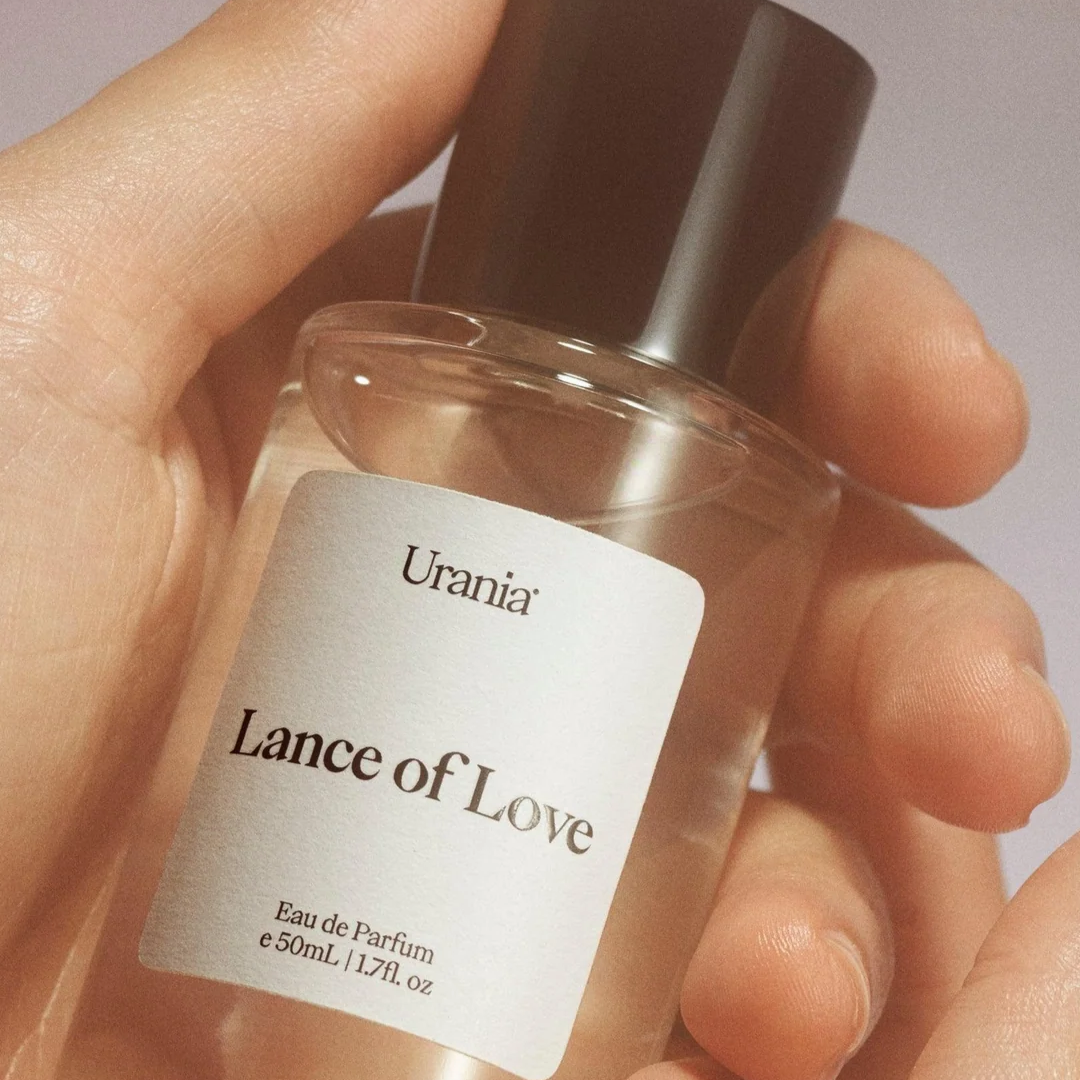 Hand holding a perfume bottle labeled 'Lance of Love' by 'Urania' against a neutral background