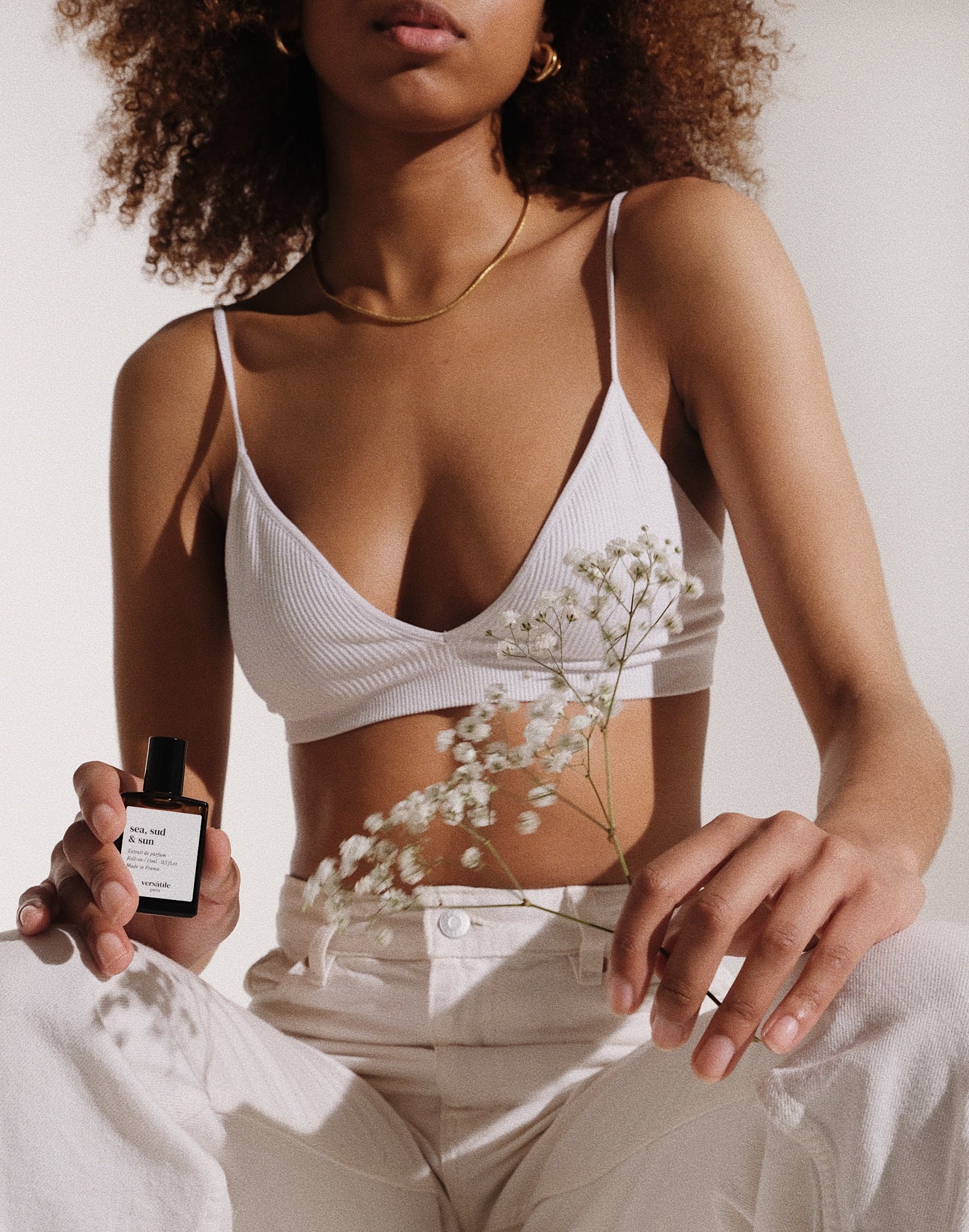 Person wearing a white bralette holding a bottle of perfume and a small plant.