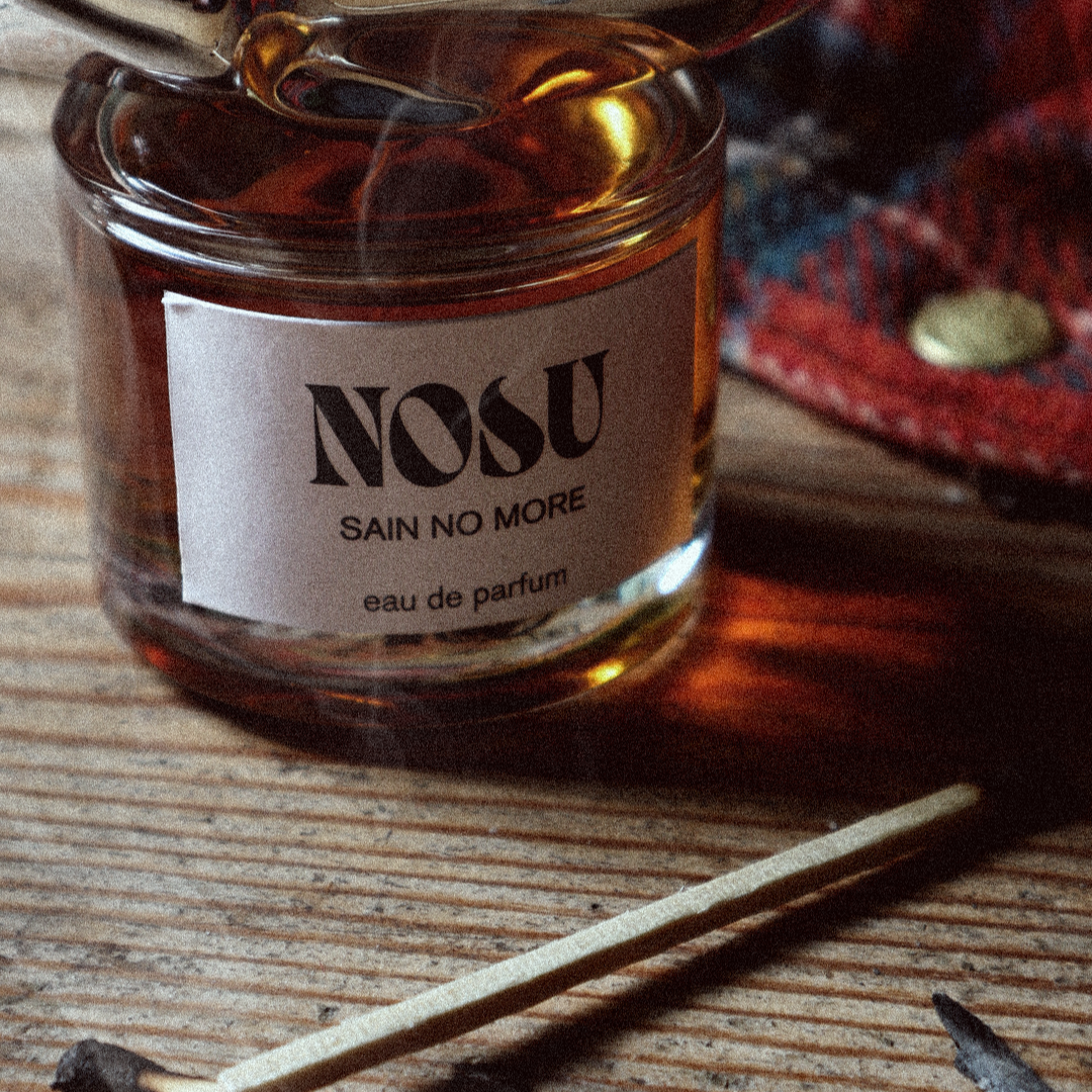 Bottle of 'NOSU Sain No More' eau de parfum with smoky matches on a wooden shack floor